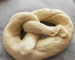 Step <p>Divida la masa en 10 partes iguales. Extienda cada parte sobre una superficie enharinada hasta formar un cordón de aprox. 60 cm cuyos extremos queden más finos que el interior. Para formar los bretzel sujete el cordón por cada extremo, cruce los extremos entre sí dos veces formando un cordón doble retorcido y lleve las puntas separadas al centro del cordón.</p> illustration